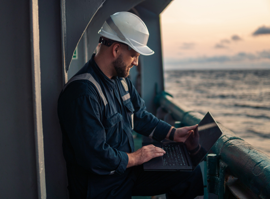 En mann med arbeidshjelp på står på et skip. Havet synes i bakgrunn og han smiler ned mot en  PC-skjerm