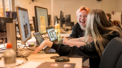 To studenter sitter ved en arbeidsstasjon med flere dataskjermer i et kontormiljø.