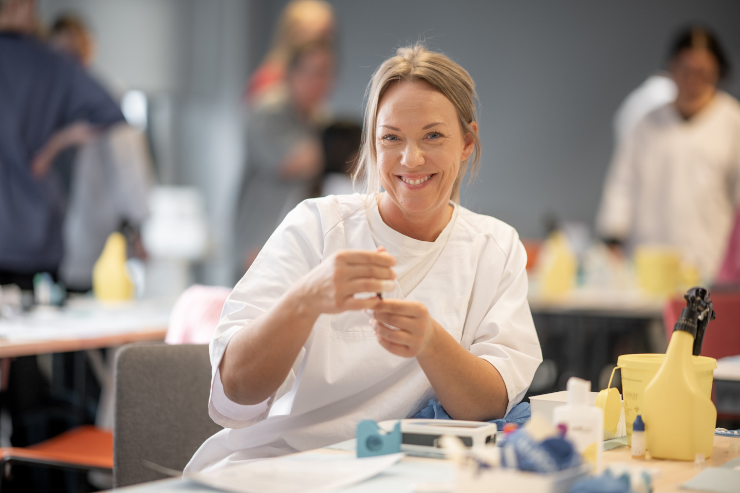 Person i hvit arbeidsuniform deltar i en praktisk øvelse og håndterer medisinsk utstyr ved et bord og smiler inn i kamera