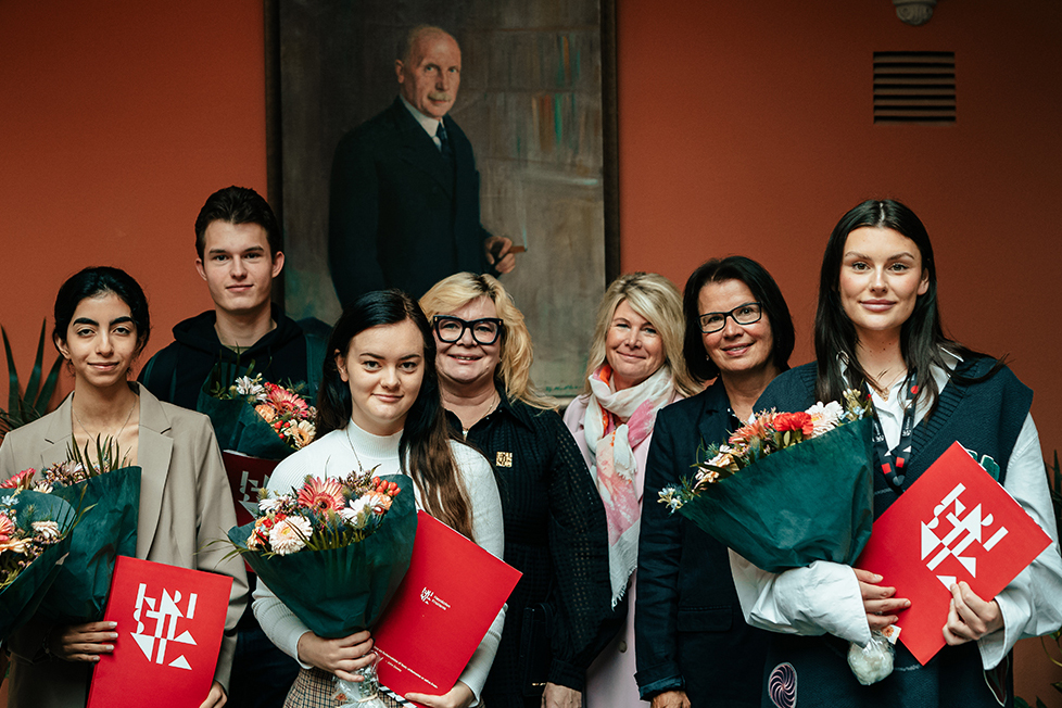 Foto av mennesker med blomsterbuketter og diplomer foran et maleri.