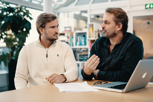 En gutt hvit genser og en mann i svart genser diskuterer sammen medhverandre mens de sitter ved et bord