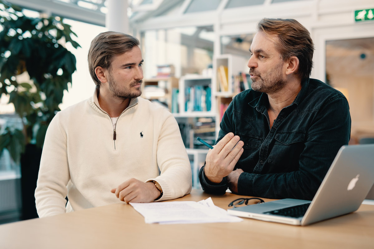 En gutt hvit genser og en mann i svart genser diskuterer sammen medhverandre mens de sitter ved et bord