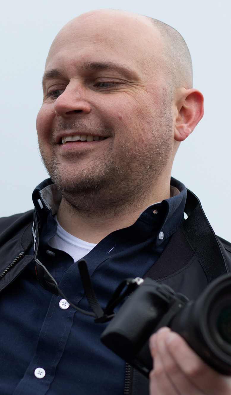 Christian Johannessen har studert digital innholdsproduksjon på fagskolen Kristiania.  Han holder  et kamera i hånden og smiler mens han ser ned. Han er skallat og har på seg en brå skjorte og svart jalle. 