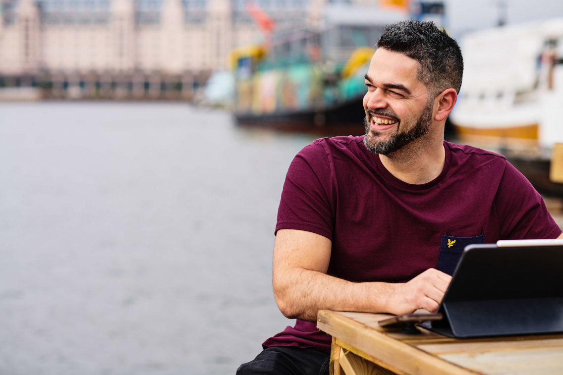 Mann med kort hår og skjegg i burgunder t-skjorte og laptop foran seg smiler.