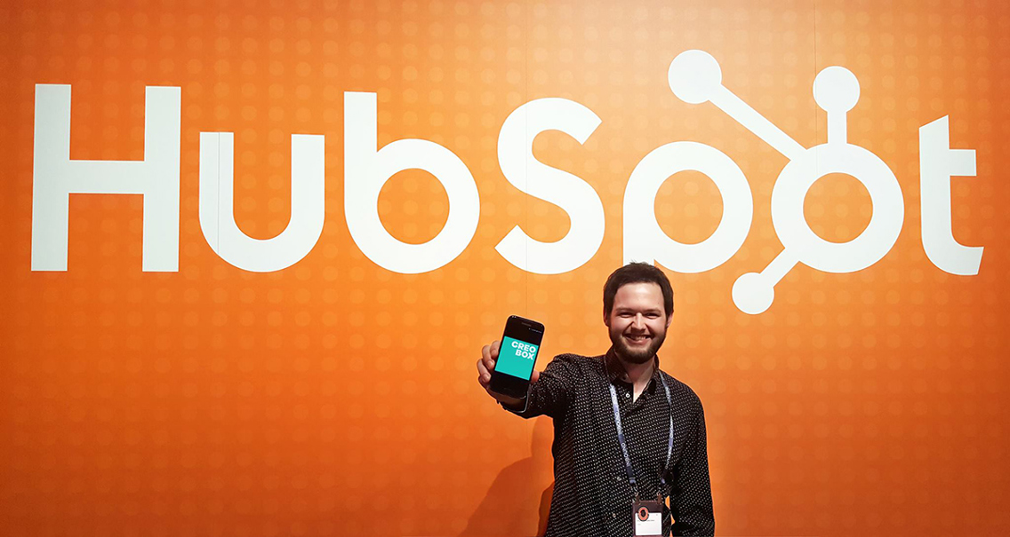Tidligere student Christopher står med den ene hånden frem mot kamera. I den holder han en smarttelefon. I bakgrunn ser vi en stor oransj vegg med logoen "Hubspot". 
