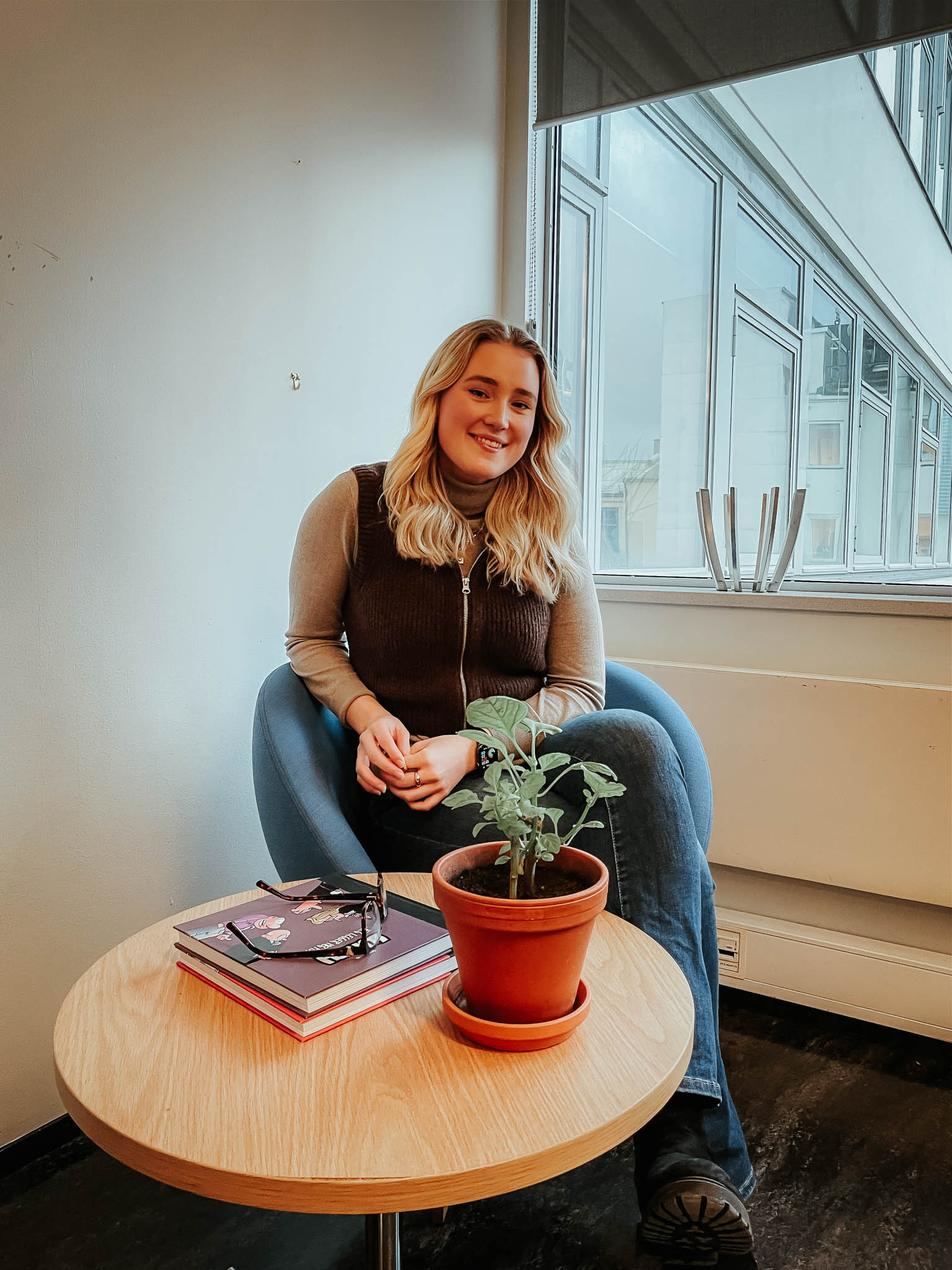 Selma Vedum Håland sitter i en turkis stol mens hun smiler mot kamera. Hun har hendene foldet til den ene siden og på bordet foran henne står det en liten potteplante. Hun har på seg strikkevest og jeans. Foto.