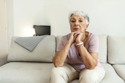 En eldre kvinne sitter i sofaen og har hodet på hendene og ser rett inn i kamera