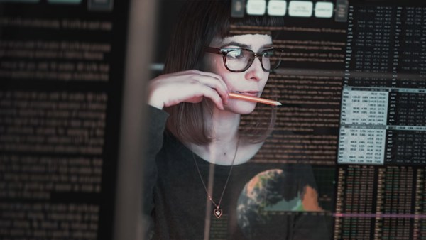 Kvinne med briller som sitter med en blyant i munnen og ser på skjermer med masse kode på 