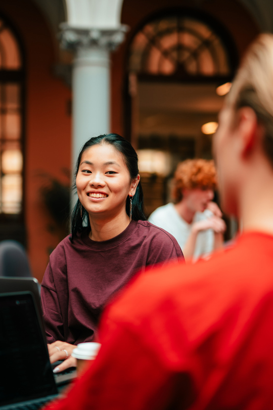 Flere studenter sitter med laptoper foran seg. En smilende kvinnelig student er i fokus. Hun sitter med fingrene på tastaturet.