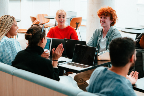 Fem studenter sitter rundt et bord med laptoper foran seg. De ser ut til å diskutere noe.
