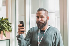 Foto av en mann med skjegg som holder opp en telefon og smiler til kamera.