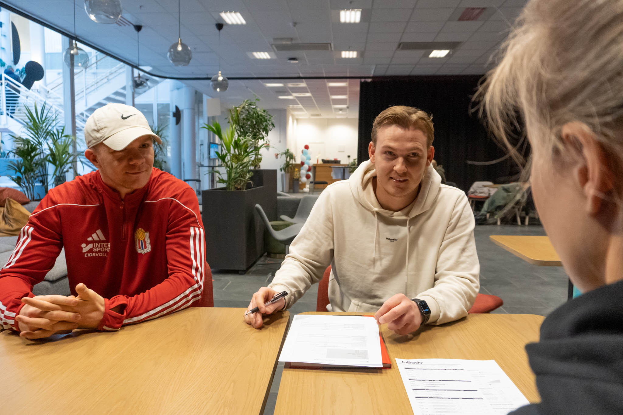Tre mennesker sitter rundt et bord mens de diskuterer deltagerens fysiske form og noterer på papir. Foto.