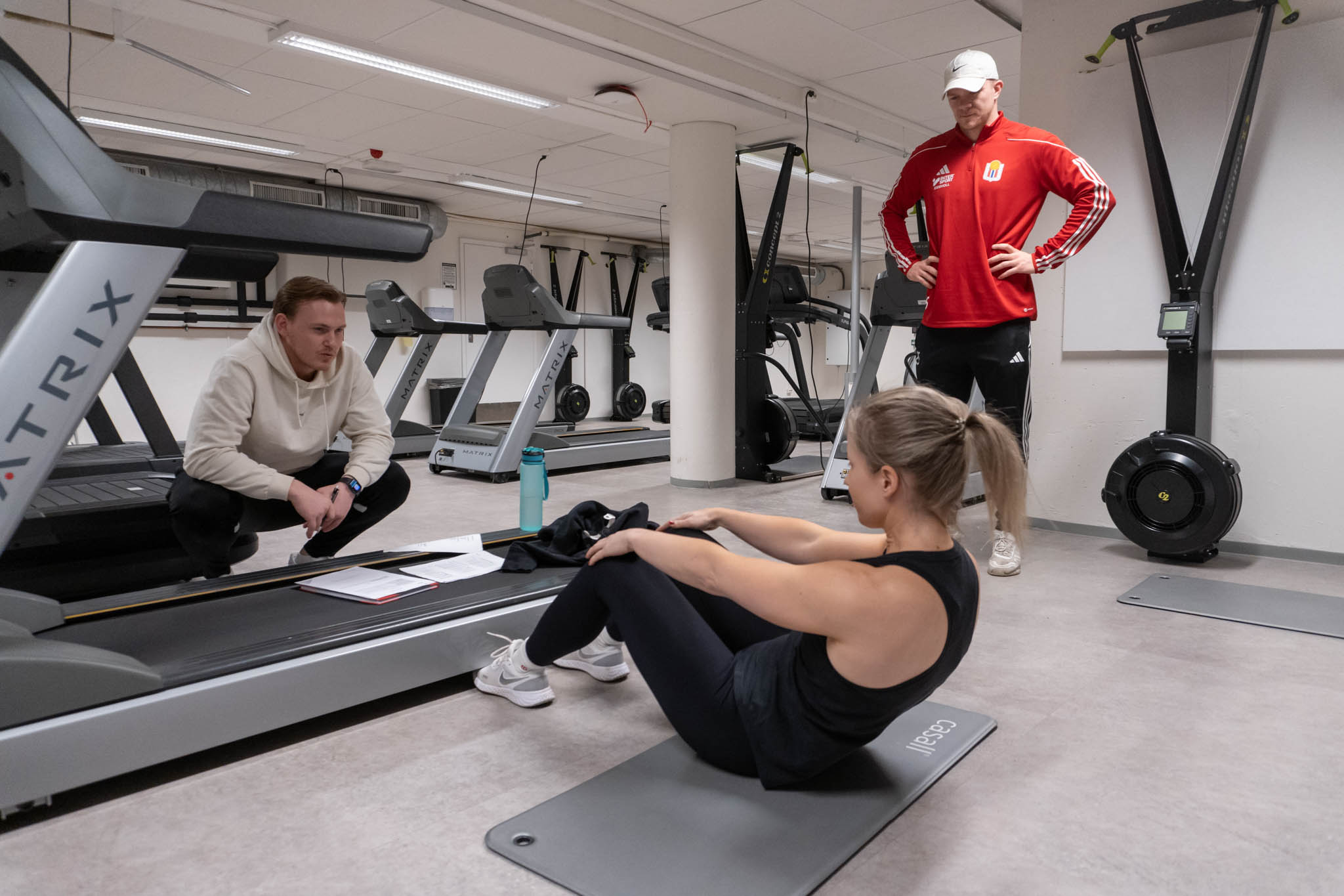 To studenter ser på mens deltager i treningsstudio tar sit ups på gulvet. Foto..