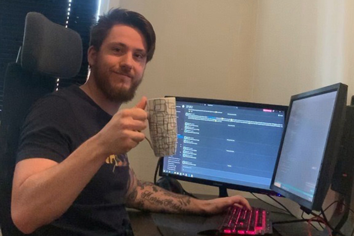 Foto av studenten ved Fagskolen Kristiania, Marius Aleksander Sletten, sitter foran en pc med en kopp kaffe og smiler fra hjemmekontoret sitt.