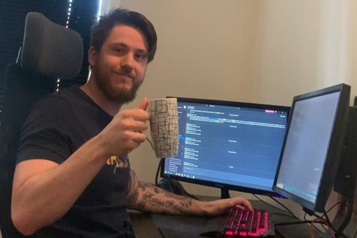 Foto av studenten ved Fagskolen Kristiania, Marius Aleksander Sletten, sitter foran en pc med en kopp kaffe og smiler fra hjemmekontoret sitt.