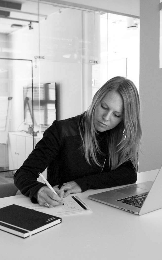 Kristine Blesvik Andersen er utdannet grafisk designer ved Fagskolen Kristiania. På bildet sitter hun ved et bord med Mac-en foran seg, og tar notater på et ark.