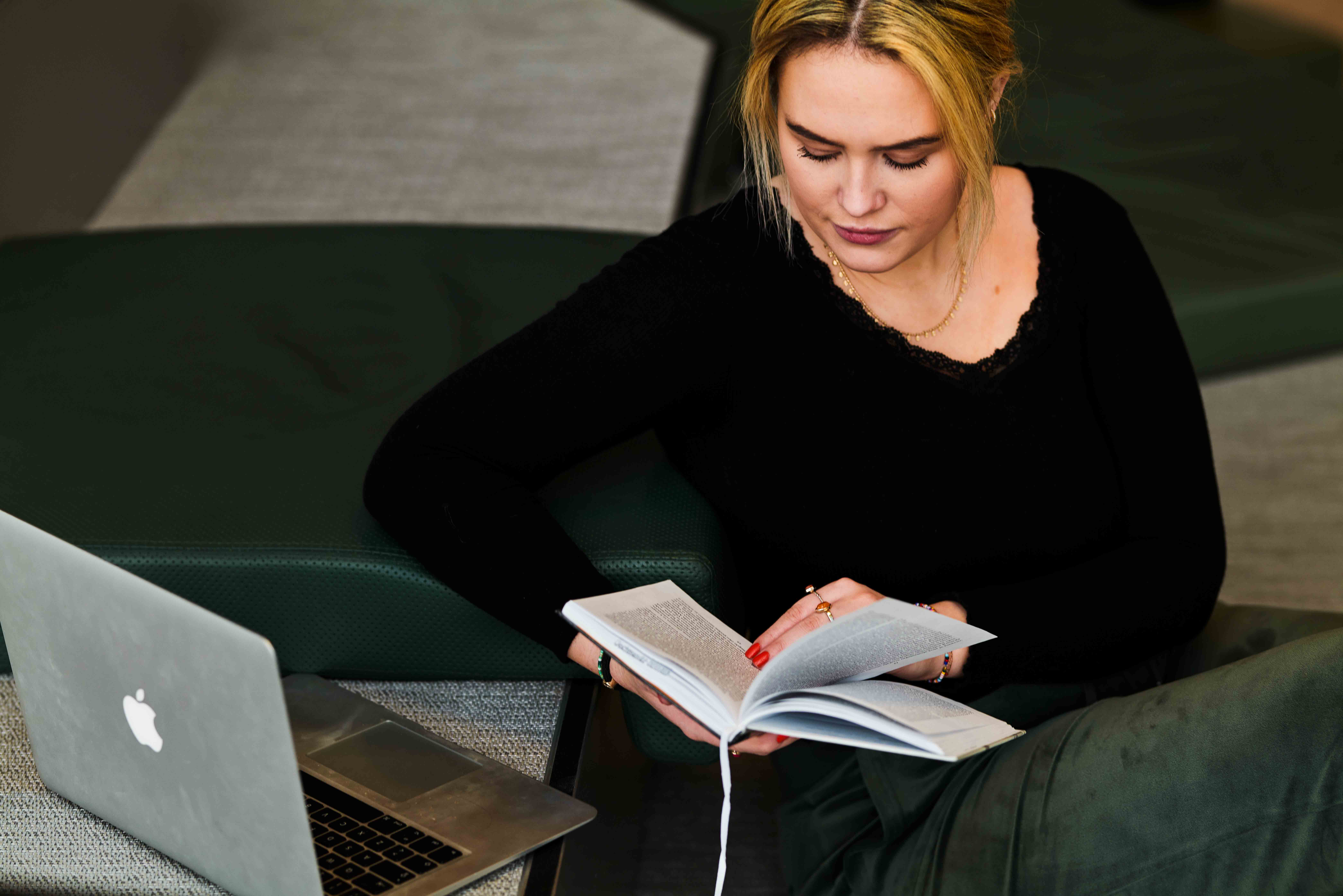 Ung blond kvinne i mørk genser leser bok ved siden av en laptop