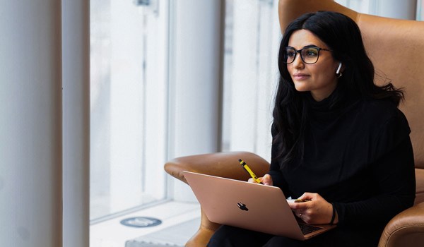 Ung kvinne med mørkt hår og briller sitter i skinnstol ved vinduet med laptop
