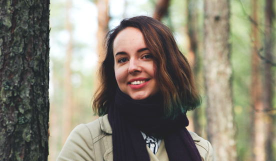 Cathrine Sandmæl er illustratør og har studert på Fagskolen Kristiania. Hun er i skogen og smiler mot kamera iført svart sjerf og beige jakke. Hun har brunt halvlangt hår. 