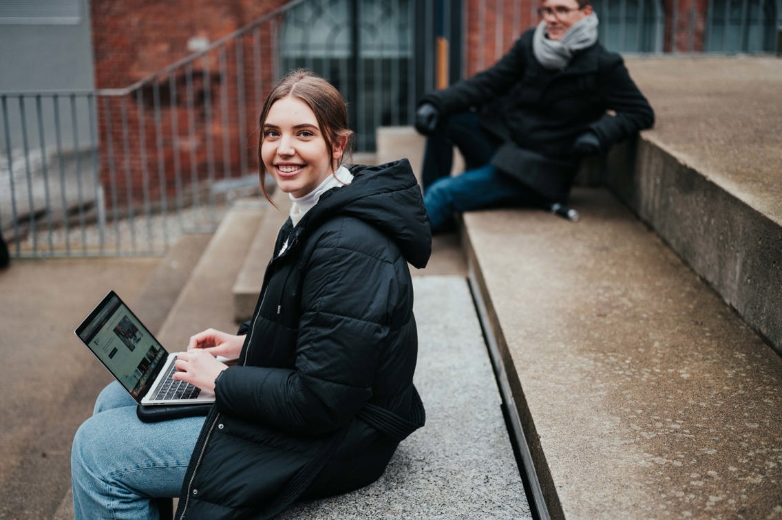 Dame studerer på laptop utendørs i høstjakke.