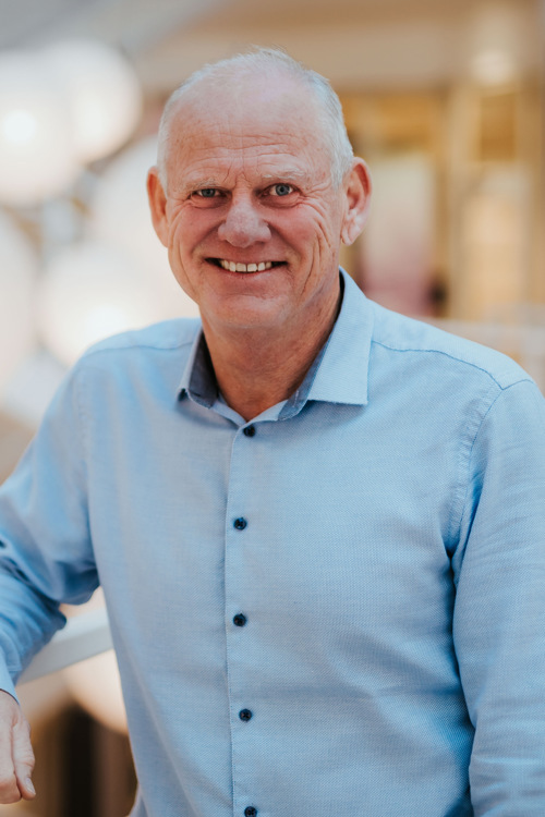 Portrett av Nils i blå skjorte, smilende til kamera mens han lener seg på rekkverket.