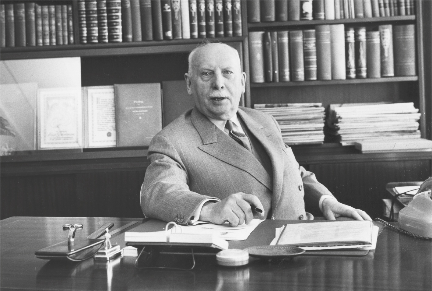Foto i sort hvitt av mann i dress som sitter ved en pult i et arbeidsrom.