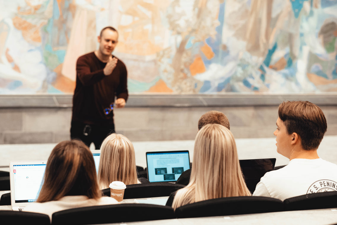 Foto av studenter i en forelesning vendt mot lærer som gestikulerer