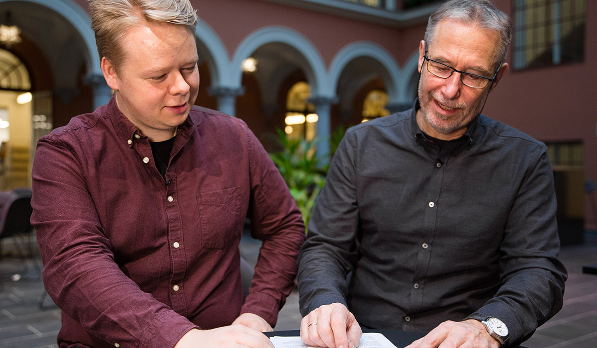 Jo Anders Alm Thoen har på seg en burgunderfarget skjorte og sitter ved siden av Tore Fagernes i en sort skjorte. Sammen ser de på papirer liggende på bordet foran seg. 