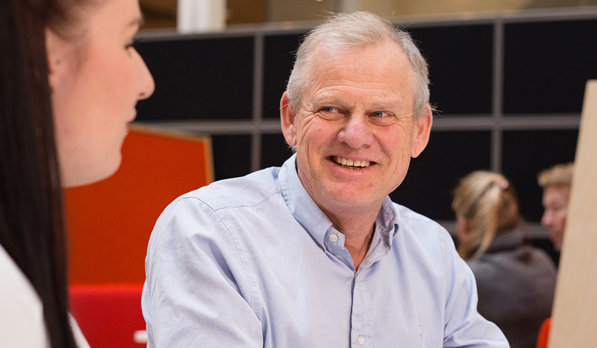Foreleser Nils Høgevold har på seg en lys skjorte og smiler mens han ser på en kvinnelig student han veileder. De sitter ved et bord og ser på hverandre.