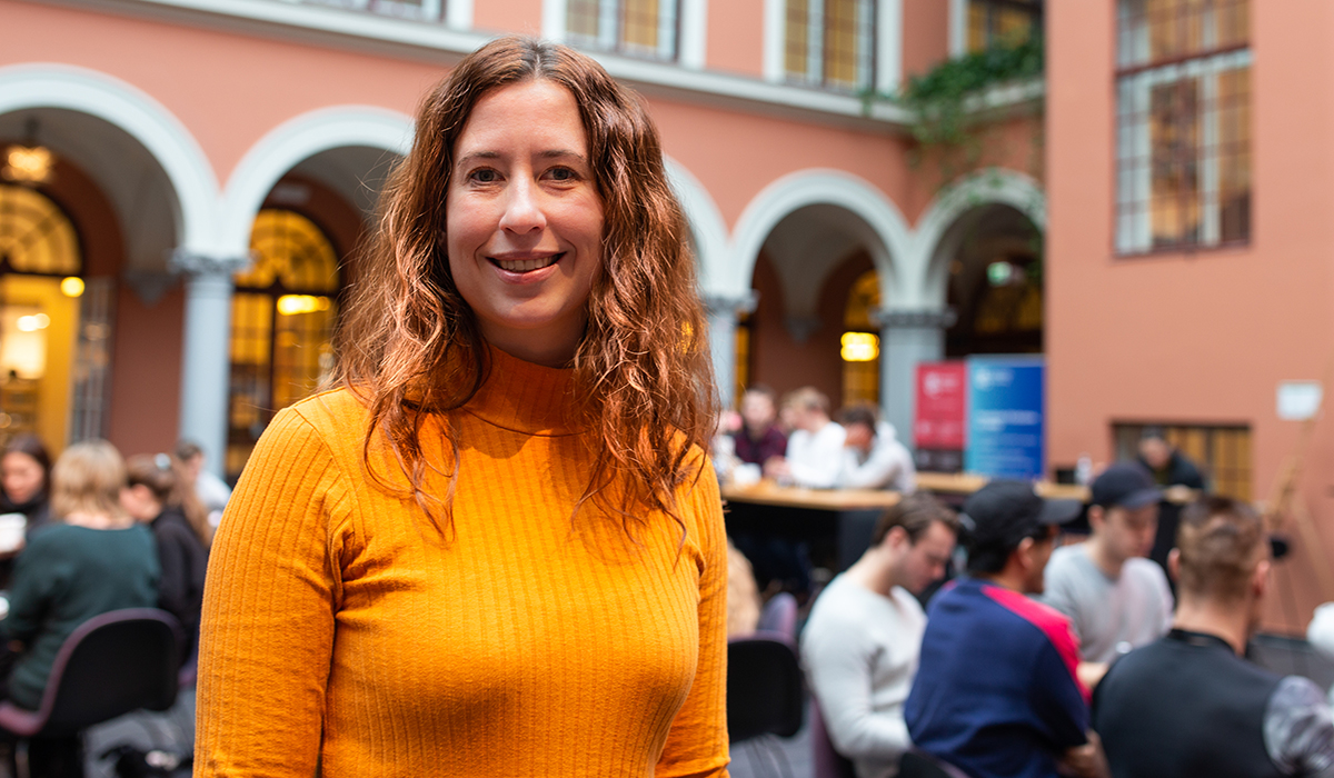 Foreleser Ragnhild Eg har rødt, krøllete hår, en oransje topp og står blant studenter og bord og smiler inn i kamera.