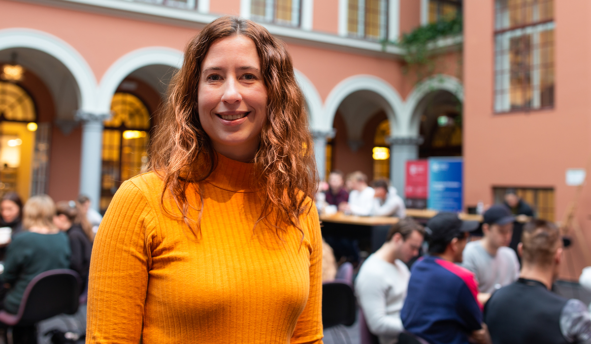 Foreleser Ragnhild Eg har rødt, krøllete hår, en oransje topp og står blant studenter og bord og smiler inn i kamera. 