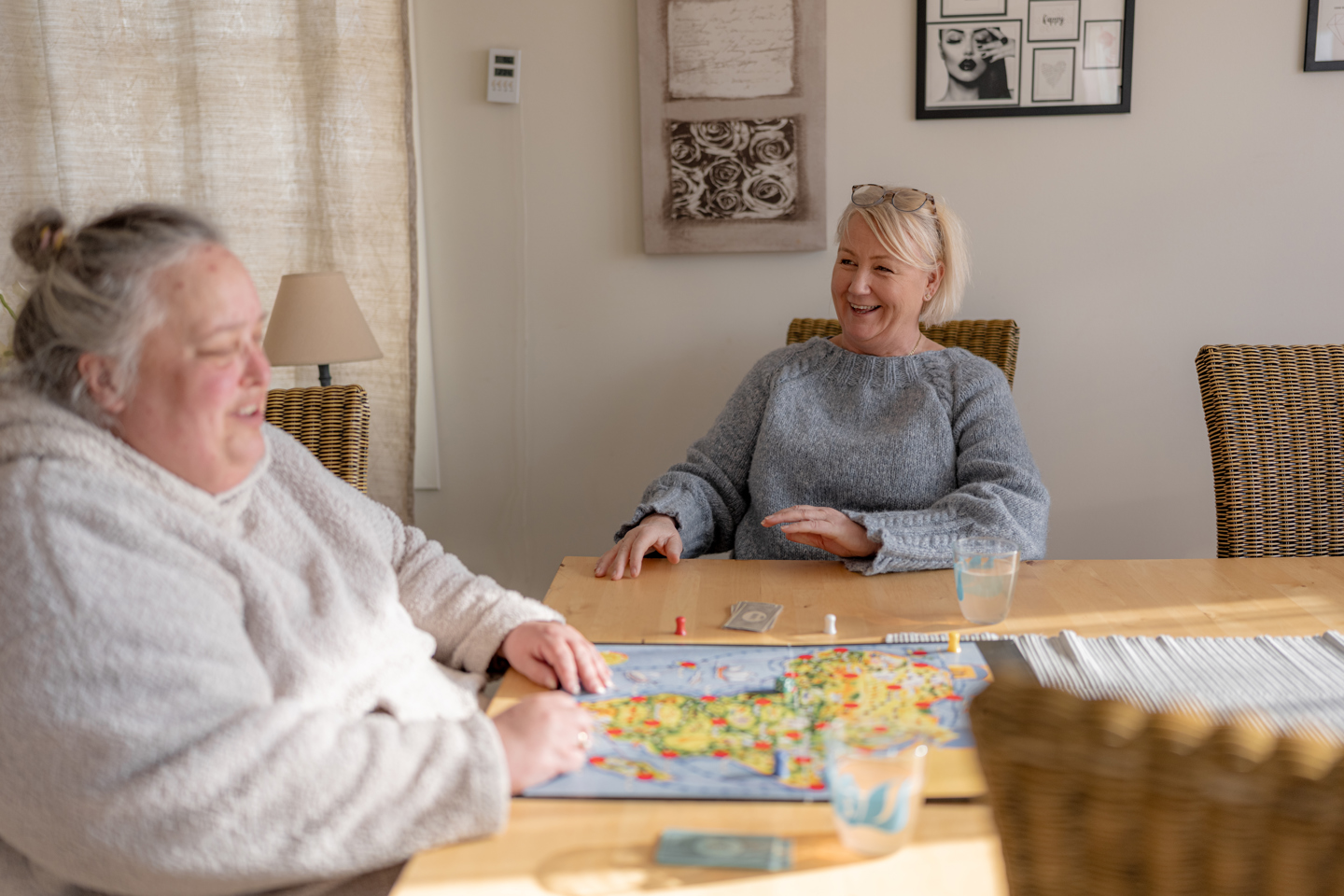 To personer sitter ved et bord og spiller et brettspill i en lys og hjemmekoselig stue.