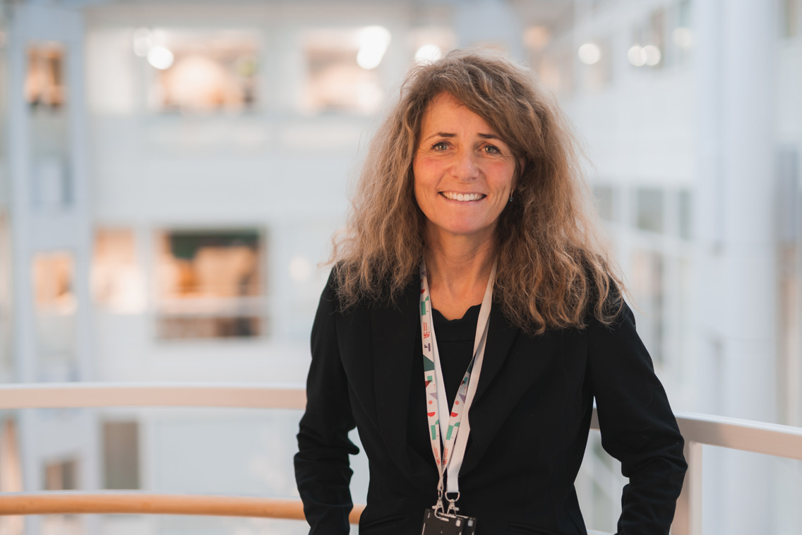 Helene ser inn i kamera med smil. Hun står foran et åpent atrium som er hvitt. 