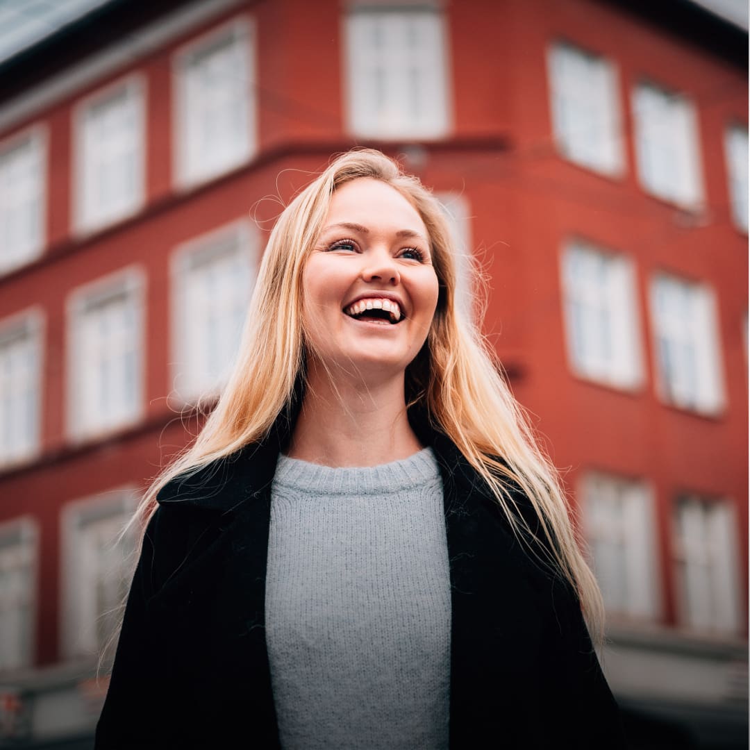 En blid jente med blondt hår står foran et rødt bygg på Grünerløkka i Oslo. 