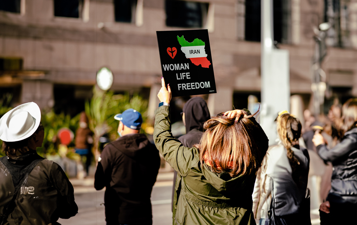 Kvinne holder plakat med ordene Woman Life Freedom og et bilde av Iran på. Hun har på seg grønn jakke og står med noen andre foran et grått bygg, tilsynelatende i protest.
