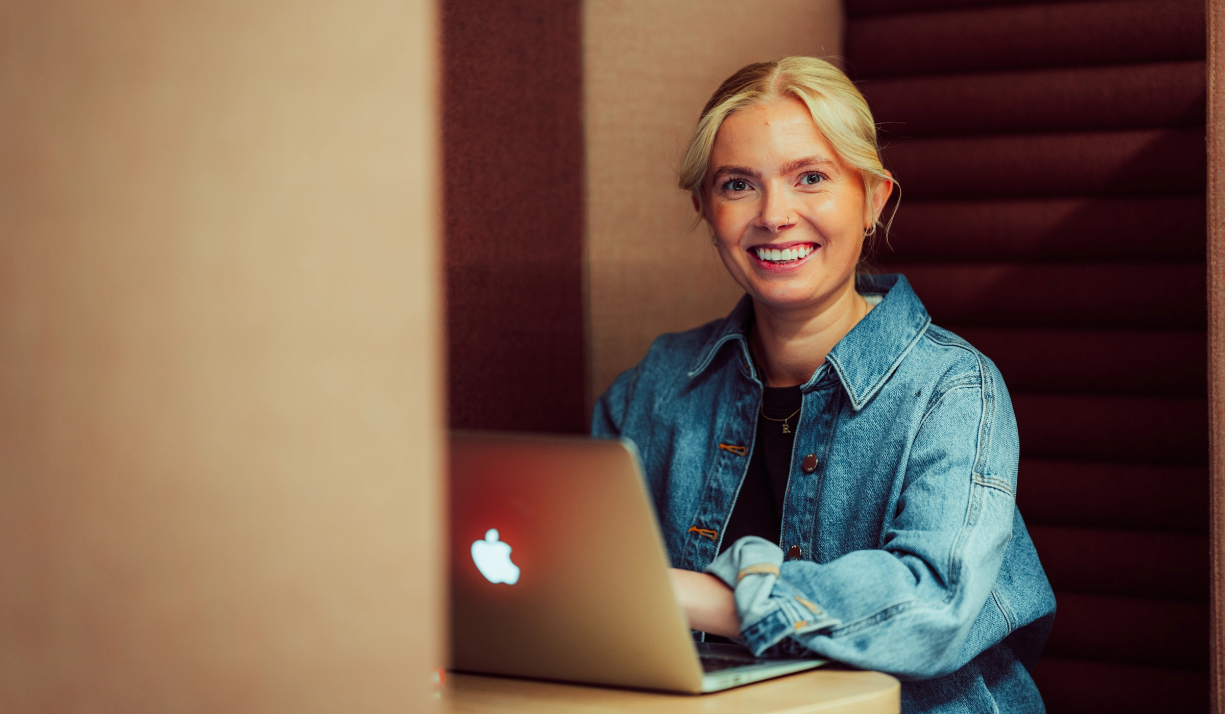 Jente sitter og jobber på en mac. Hun smiler til kamera.