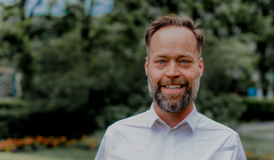 Mann i hvit skjorte smiler ute i en park.