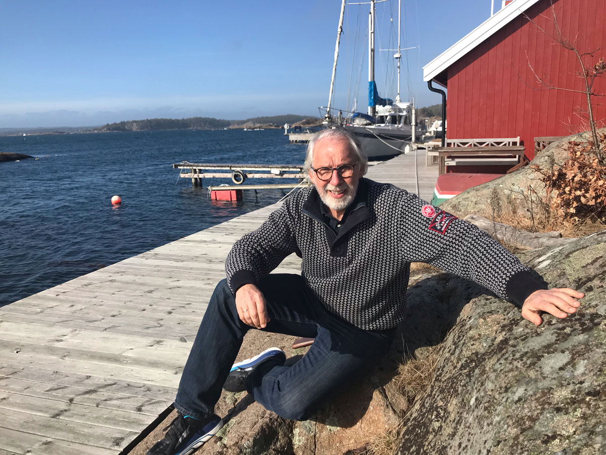 Carl-Erik Grimstad på brygge i fritidsklær med sjøhus og vann bak seg.