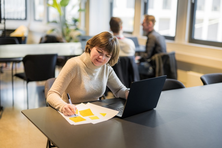 Person sitter ved et bord og arbeider med notater og bærbar PC i et undervisnings- eller kontormiljø.