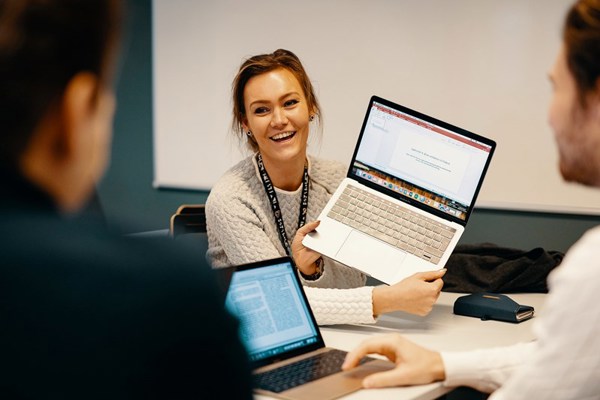 Foto av kvinnelig student som holder opp en laptop for å vise til to mannlige medstudenter.