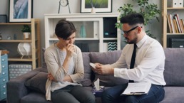 A woman sits on a sofa looking upset while a man is handing her a tissue