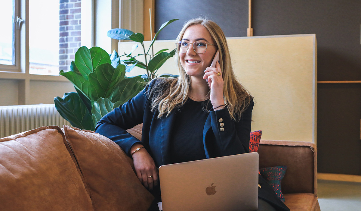 En dame med langt blondt hår sitter i en brun skinnsofa og snakker i telefonen. med en Mac på fanget. Hun har på seg en mørkeblå dressjakke og briller.