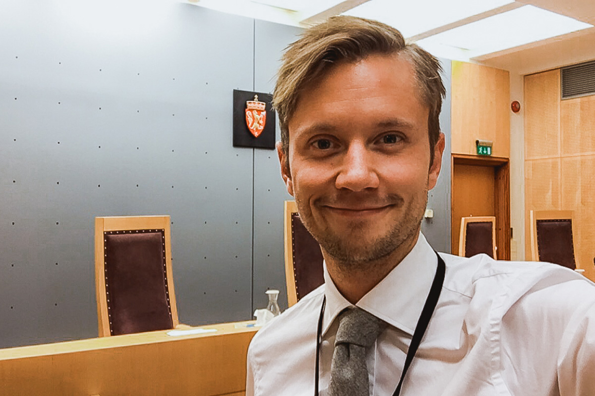 Patrick Neumann står i et rettslokale. Han har på seg en hvit skjorte og et grått slips. Han har kort blondt hår, skjegg og smiler til kamera.