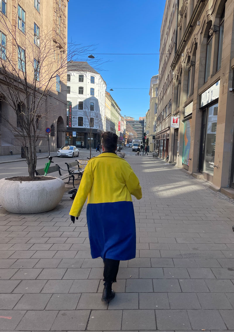 Dame med kåpe i Ukrainas farger går nedover gaten med knall blå himmel.