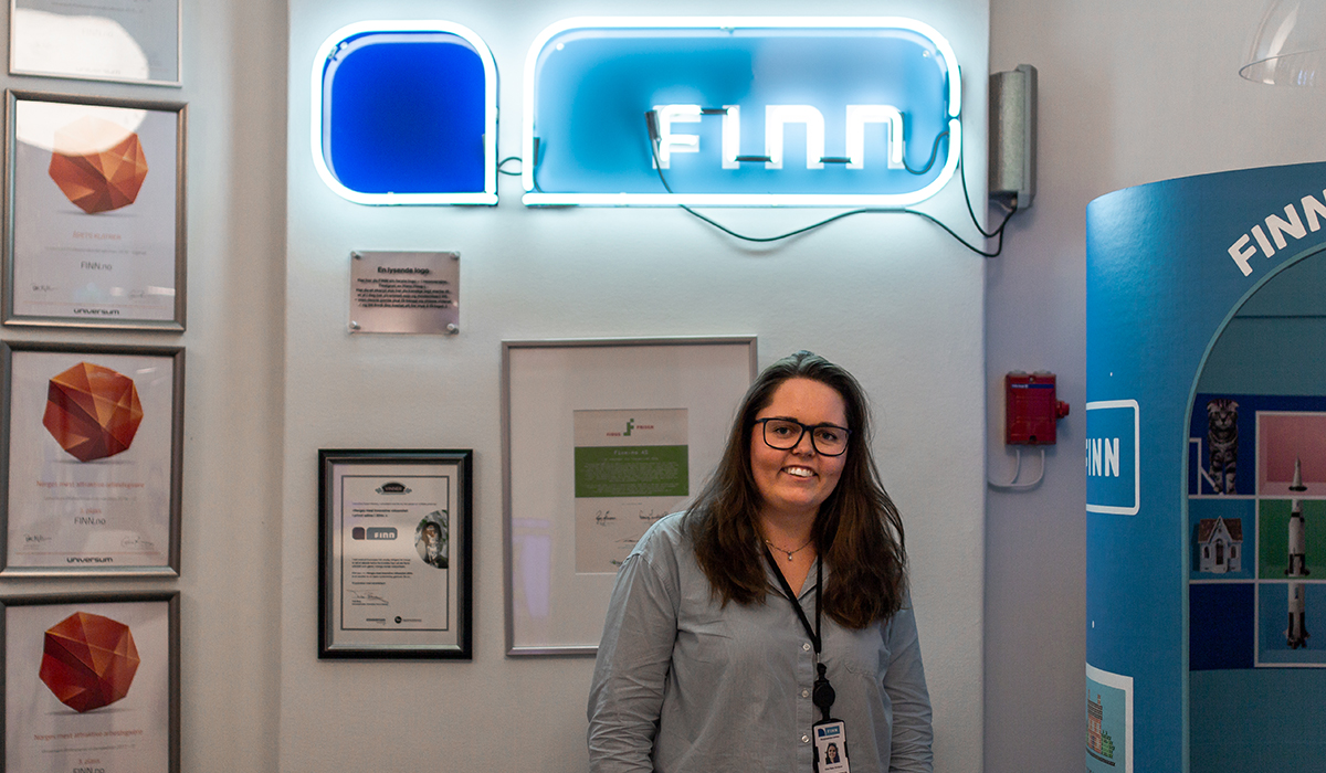 Kvinne foran vegg med diplomer og en Finn-logo med neonlys.