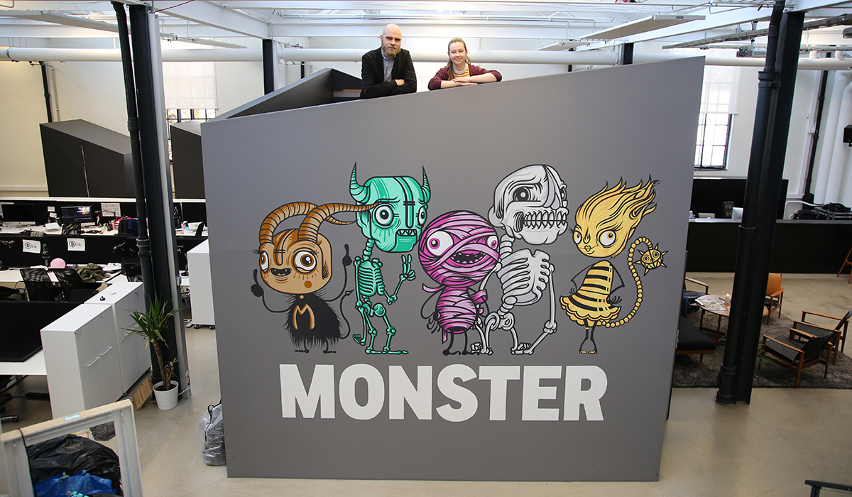 Embla Bugge og Anders Bergersen står på toppen av en diger boks med Monster-logo på inne på Monster-kontoret