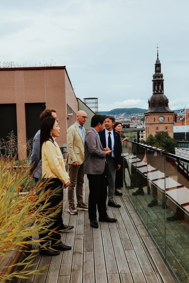 Japanese delegation on the rooftop terrace of Kristiania University of Applied Sciences, with Oslo Cathedral in the background.