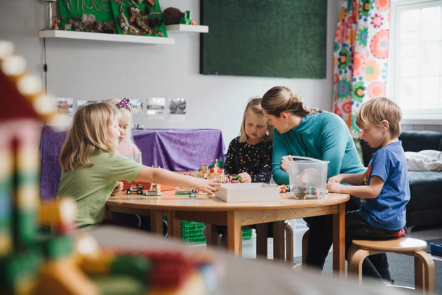 En voksen bøyer seg for å hjelpe tre barn som leker med byggeklosser ved et bord i et klasserom med fargerike dekorasjoner.