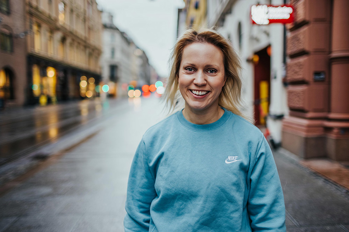 Kamilla Kolvig i blå genser og blondt hår i prinsens gate i Oslo, omgitt av høye, gamle bygninger på hver side.