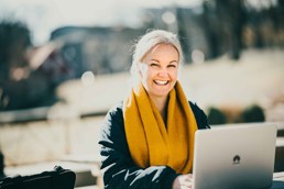 Foto av kvinne med gult skjerf som sitter med laptop på en benk i en park.